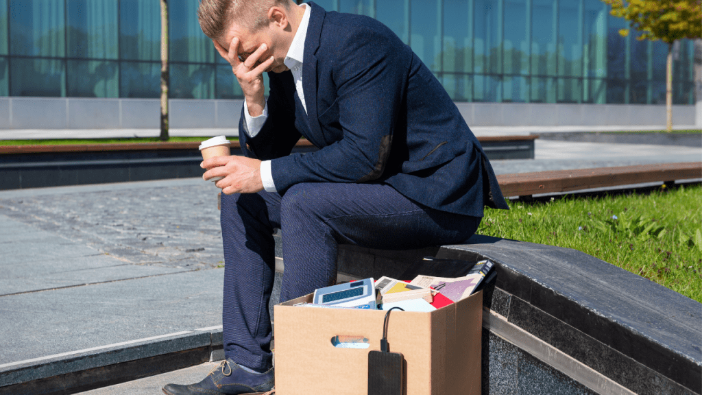A man sitting outside an office building with a box of belongings after job loss, representing the stress of losing employer health coverage and the need for short term insurance solutions